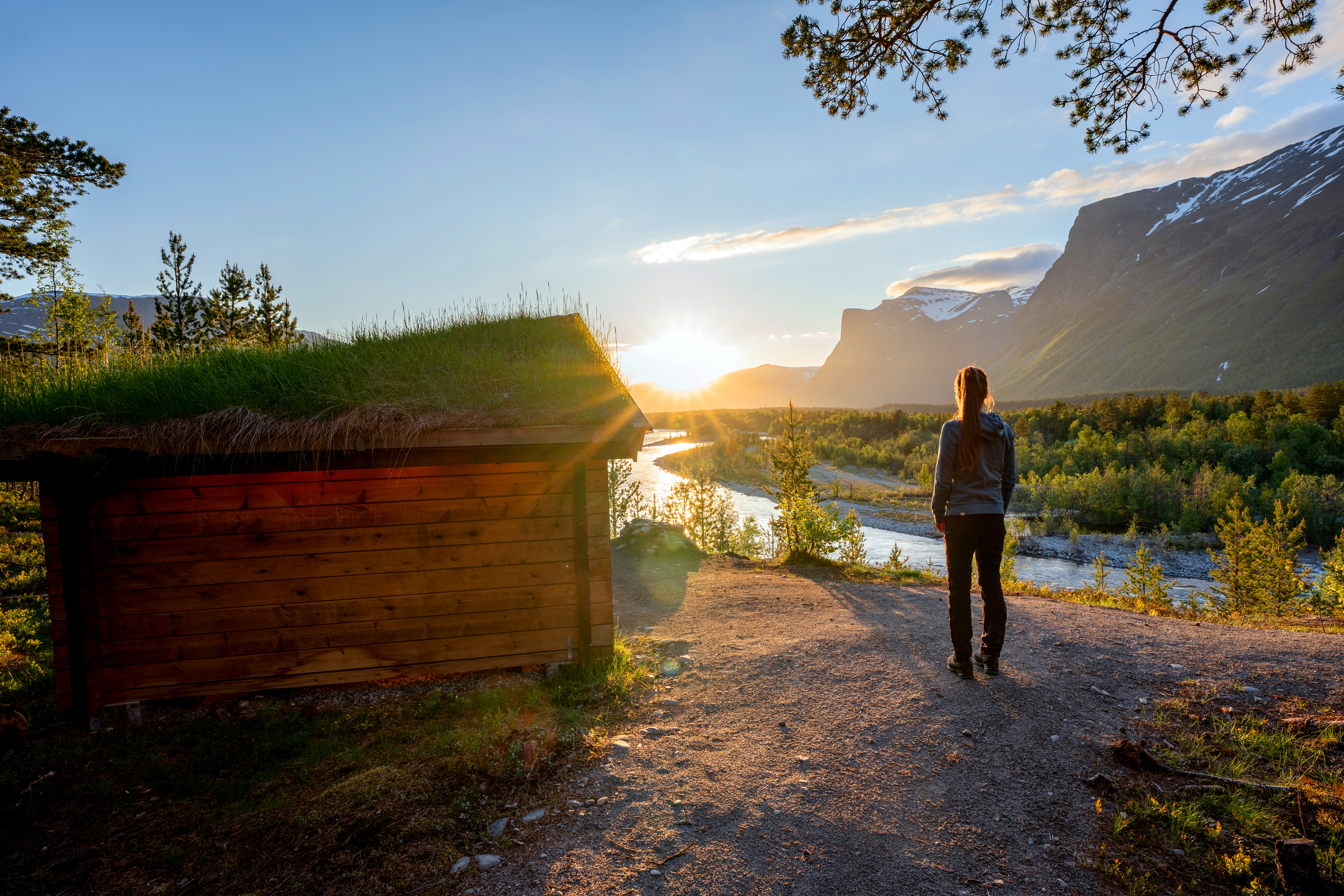 Person st&aring;r p&aring; sti i solnedgang over elv og fjell.