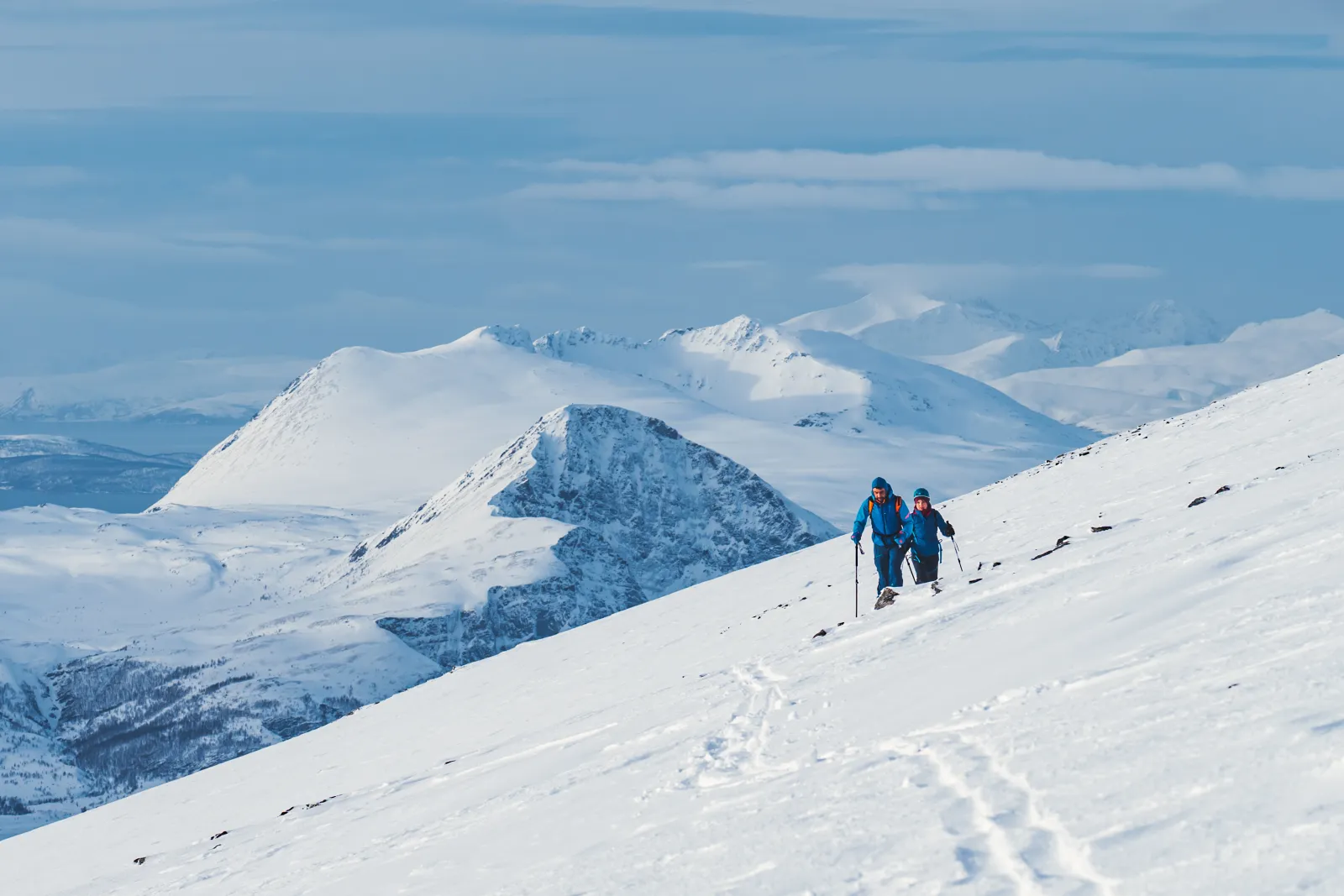 To skil&oslash;pere p&aring; vei opp en sn&oslash;dekt fjellside med flere fjelltopper i bakgrunnen.