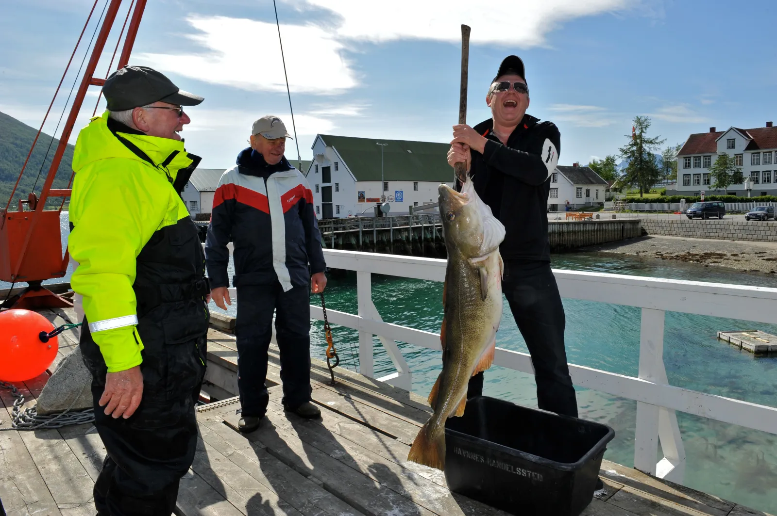 Person viser frem en stor torsk p&aring; en brygge, mens andre ser p&aring;, med bebyggelse i bakgrunnen.