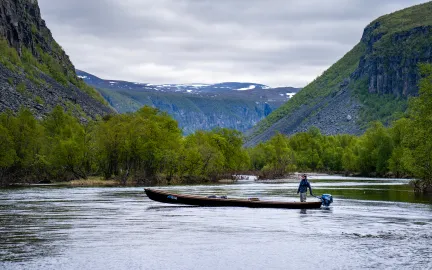 Person st&aring;r i b&aring;t p&aring; elv omgitt av dal og fjellandskap.