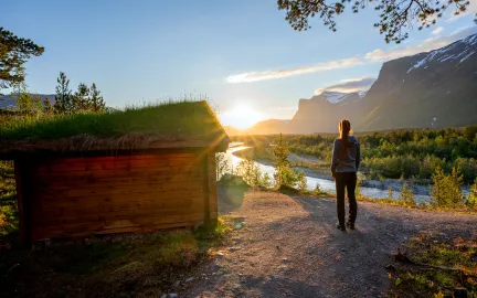 Person st&aring;r p&aring; sti i solnedgang over elv og fjell.