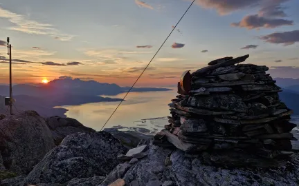 Solnedgang over fjord og fjell, sett fra en fjelltopp med en stor steinvarde i forgrunnen.