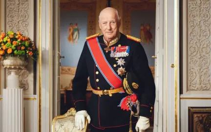 H. M Kong Harald V 1937-2026 Kong Harald står i gallauniform med ordener og medaljer i et rikt utsmykket rom på Slottet. Han holder en sabel i den ene hånden og støtter den andre på en stol.
