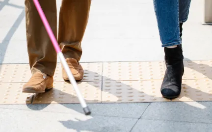 En person med hvit stokk g&aring;r sammen med en annen person over taktil merking.