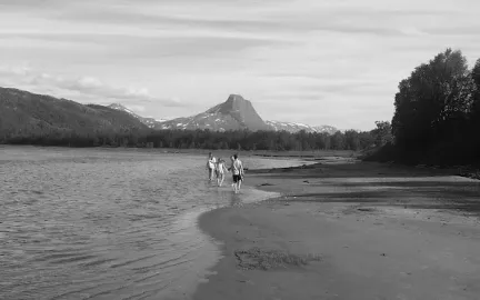 Utsikt over Kv&aelig;nnes med strand, vann og fjell i bakgrunnen. Tre personer g&aring;r langs vannkanten.