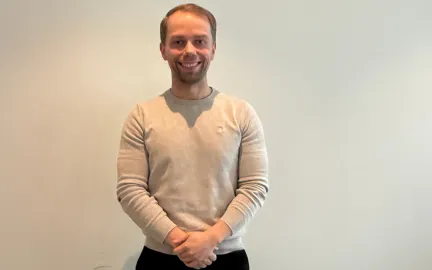 En person st&aring;r oppreist foran en lys vegg med hendene samlet foran kroppen. Personen har p&aring; seg en lys gr&aring; genser og m&oslash;rke bukser. Gulvet er m&oslash;rkt, og bildet er tatt innend&oslash;rs med n&oslash;ytral bakgrunn.