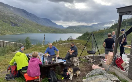 En turgruppe med hunder tar en pause ved et bord ved et fjellvann, omgitt av gr&oslash;nne fjellsider.