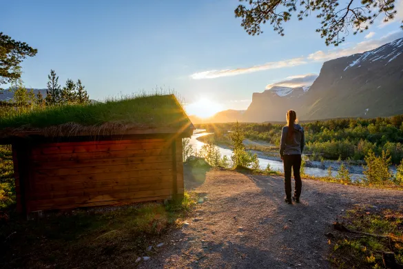 Person st&aring;r p&aring; sti i solnedgang over elv og fjell.