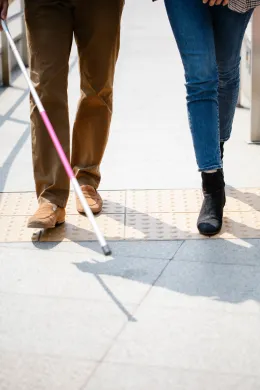 Tilgjengelighet og ledsaging En person med hvit stokk går sammen med en annen person over taktil merking.