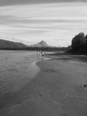 Barn på stranda på Kvænnes Utsikt over Kvænnes med strand, vann og fjell i bakgrunnen. Tre personer går langs vannkanten.