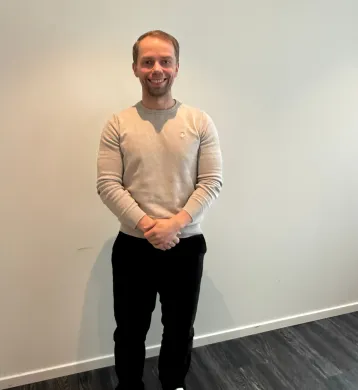 Økonomisjef Andreas Knutsen En person står oppreist foran en lys vegg med hendene samlet foran kroppen. Personen har på seg en lys grå genser og mørke bukser. Gulvet er mørkt, og bildet er tatt innendørs med nøytral bakgrunn.