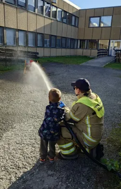 Barn får prøve brannslange t barn sprøyter vann med en brannslange mens en brannkonstabel sitter på huk og hjelper til.